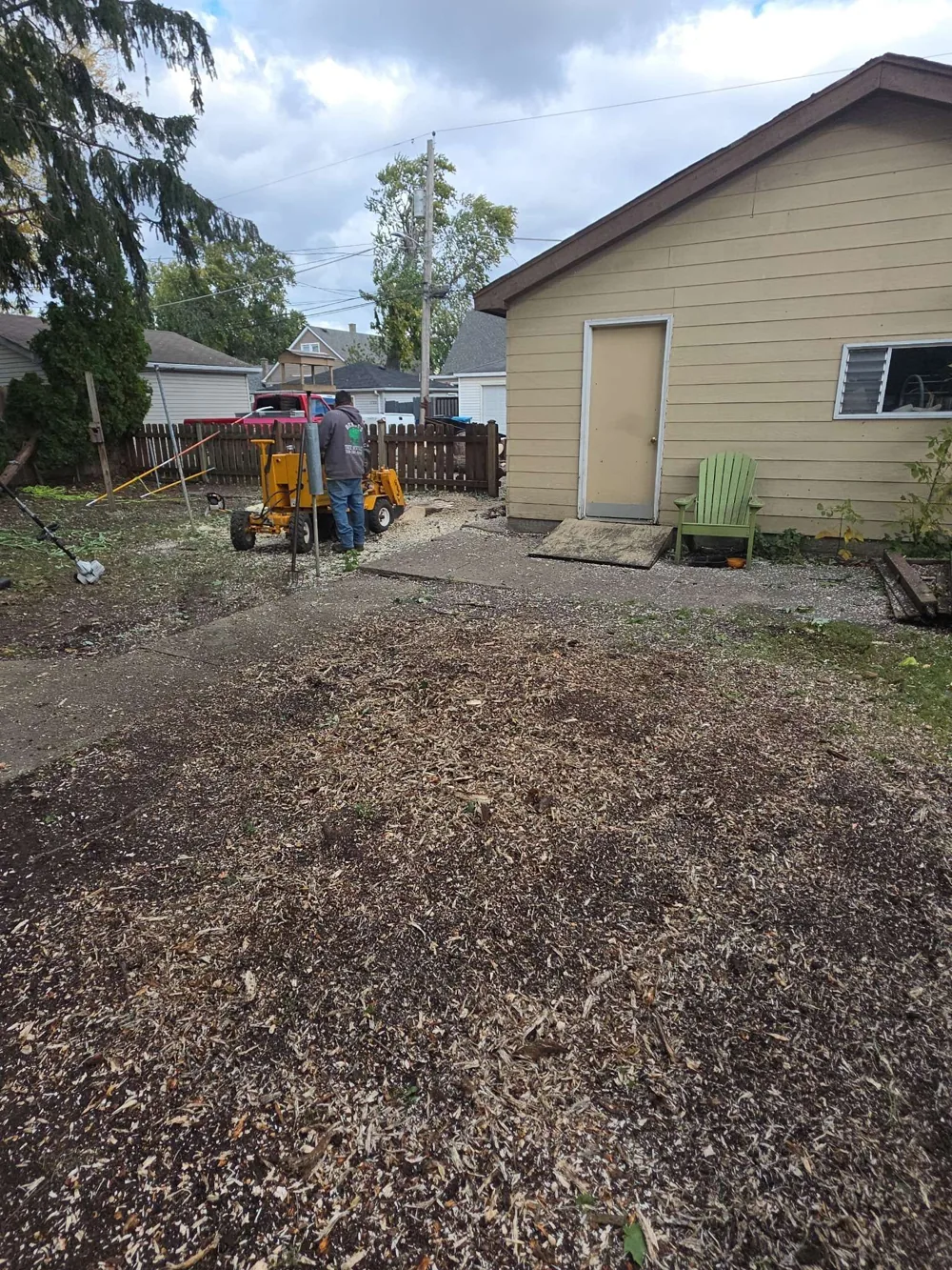 Worker with stump-removal equipment in the cleared yard beside the same garage, with wood chips on the ground.
